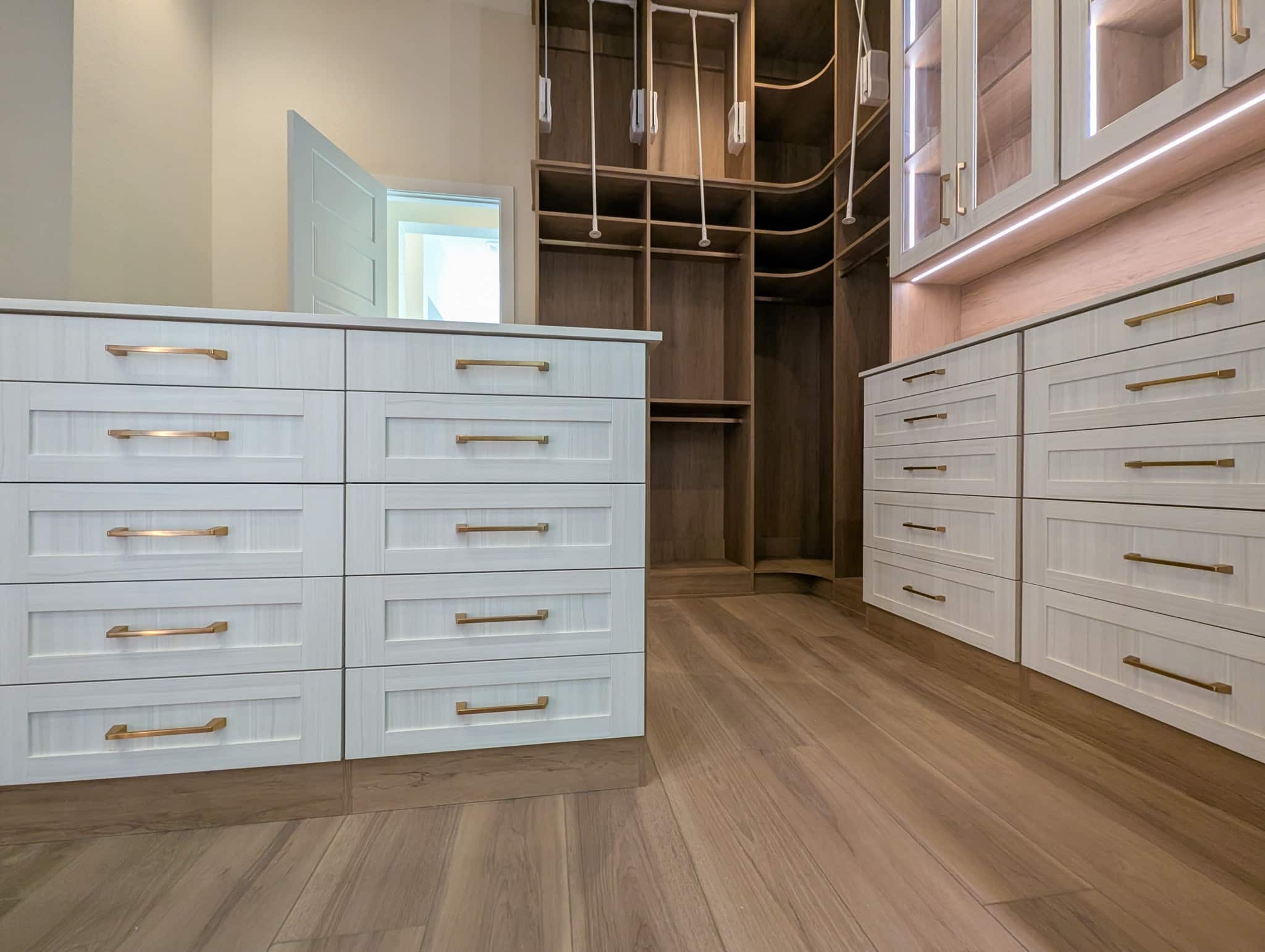 Custom walk-in closet in brown and white with island, pull down rods, shoe wall and LED lighting. Floor to ceiling shelves and glass cabinets