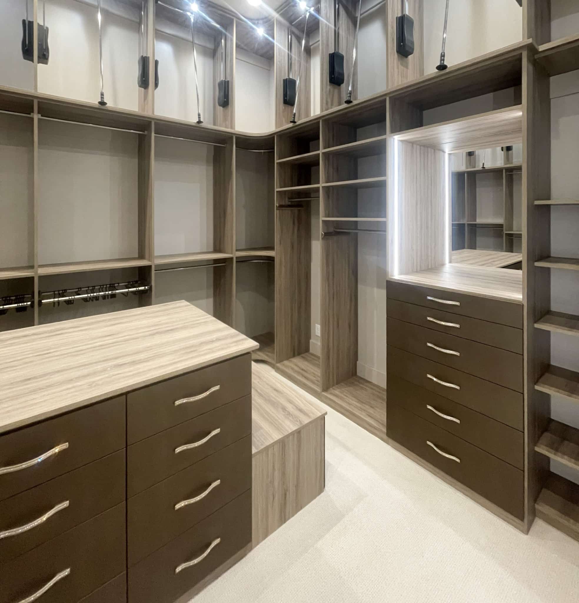 Luxury custom walk-in closet with brown textured drawers, pull down rods, LED lighting, mirror, island, and jeweled handles