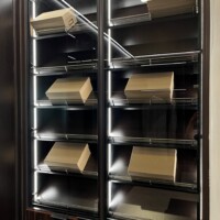 This luxury custom walk in closet features slanted shoe shelves with lighting and glass doors for easy navigation.